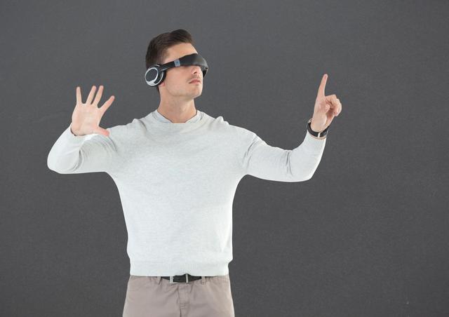 Man wearing virtual reality headset, immersed in an interactive experience, gesturing with hands against a gray background. Ideal for illustrating concepts of modern technology, innovation, and virtual reality in digital and print media. Suitable for blogs, articles, and advertisements related to VR technology, gaming, and immersive experiences.