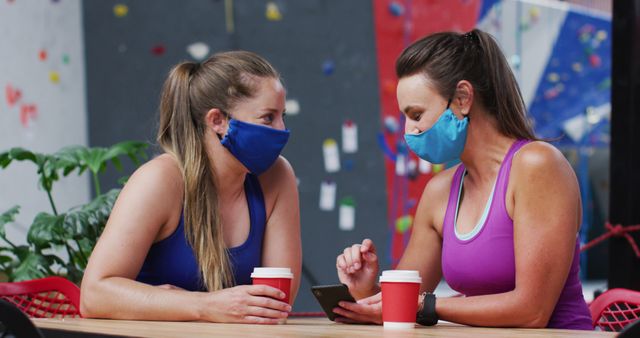 Two happy caucasian women in face masks using smartphone in cafe at indoor climbing wall. fitness and leisure during coronavirus covid 19 pandemic.