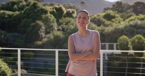 Smiling woman standing on sunlit balcony with arms crossed and casual activewear