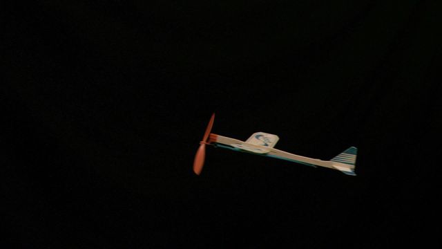 This shot captures a toy elastic propeller plane flying in slow motion against a black background. Ideal for use in educational content about basic principles of flight, presentation on motion and air dynamics, children’s entertainment materials, or creative advertisements highlighting themes of freedom, flight, and childhood nostalgia.