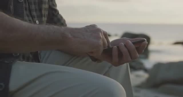 Midsection of senior Caucasian man tapping on smartphone while sitting by sea at sunset. Perfect for promoting technology use among older adults, emphasizing relaxation and outdoor activities for seniors, or evoking serene, nature-oriented backgrounds for ads or websites.