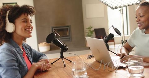 Multiethnic women recording cozy home podcast with laptops, microphones and headphones