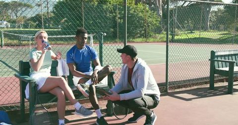 Mixed-ethnicity tennis players resting and chatting on outdoor court bench