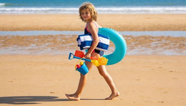 Child walking on sandy beach carrying turquoise inflatable ring, striped towel and bucket