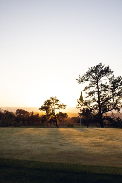 This image captures a serene golf course at sunset, with a yellow flag marking a hole amidst a grassy field. Silhouette trees stand against a clear sky, creating a peaceful and picturesque scene. Ideal for use in sports advertisements, nature-themed promotions, or as a calming background for websites and presentations.