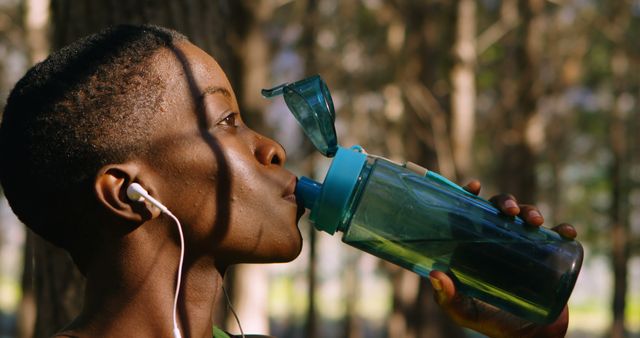 Athletic woman drinking from water bottle while exercising outdoors, earphones in ears. Picture suitable for promoting healthy lifestyle, outdoor activities, fitness products, and hydration awareness.