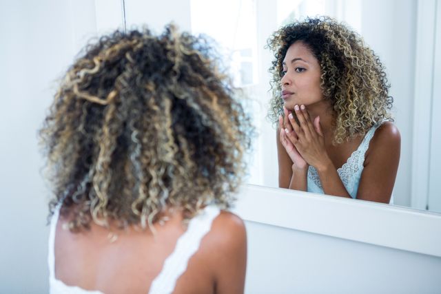 Young woman with curly hair checking her skin in the bathroom mirror. Ideal for content related to skincare routines, beauty tips, self-care practices, and personal hygiene. Can be used in articles, blogs, and advertisements focusing on beauty and health.