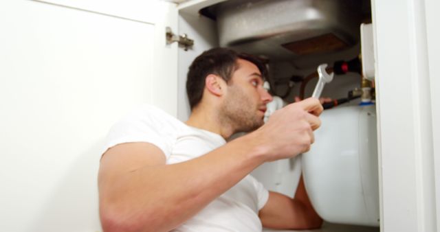 Male plumber dressed in a white t-shirt working under kitchen counter repairing sink pipes. Image is suitable for articles on home maintenance, DIY plumbing, and professional plumbing services. Can also be used in marketing materials for plumbing companies and handyman services.