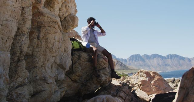 Young man sitting on rocky mountain, using binoculars to view distant landscape. Background includes rugged mountains and clear skies. Scene suits outdoor adventure, travel advertisements, nature blogs, and exploration themes.