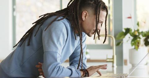 African American man leaning on kitchen counter using laptop, working remotely in minimalist home