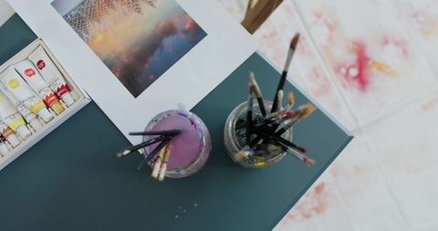 Top-down artist workspace with paintbrush jars, purple rinse water, acrylic tubes and print