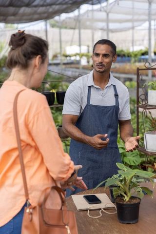 Nursery worker advising customer about potted plant care in shaded greenhouse retail space