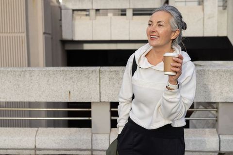 Mature woman enjoying coffee on urban parking deck wearing white hoodie backpack and watch