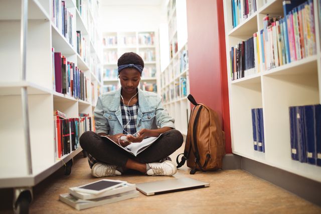 Young African American schoolgirl sitting on floor in library, listening to music on headphones while studying. Surrounded by books, backpack, tablet, and laptop, she is focused on her work. Ideal for educational content, student life, academic resources, and back-to-school promotions.