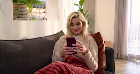 Blonde woman relaxing on sofa texting on smartphone smiling wrapped in red blanket cozy living room