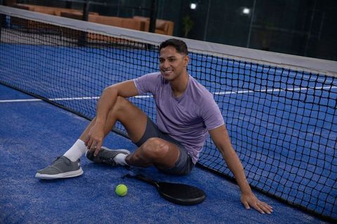 Smiling athlete sitting by net resting after padel session on blue indoor court with racket