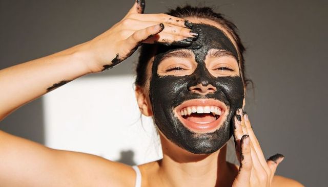 Smiling woman applying charcoal face mask at home with sunlit skin and smudged fingers