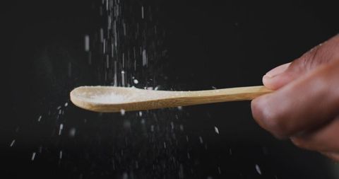 A hand elegantly sprinkles salt from a wooden spoon, grains suspended in mid-air against a black background. This image captures the art of seasoning, emphasizing fine cooking techniques and gourmet food preparation. It is perfect for culinary blogs, recipe websites, or spice product advertisements, showcasing finesse in kitchen practices and enhancing culinary content with a stylish touch.