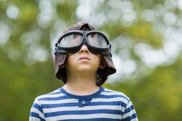 Young boy wearing aviator goggles and striped shirt, standing outdoors in park, pretending to be a pilot. Ideal for concepts of childhood imagination, creativity, outdoor play, and adventurous spirit. Suitable for educational materials, children's books, and advertisements promoting outdoor activities and imaginative play.