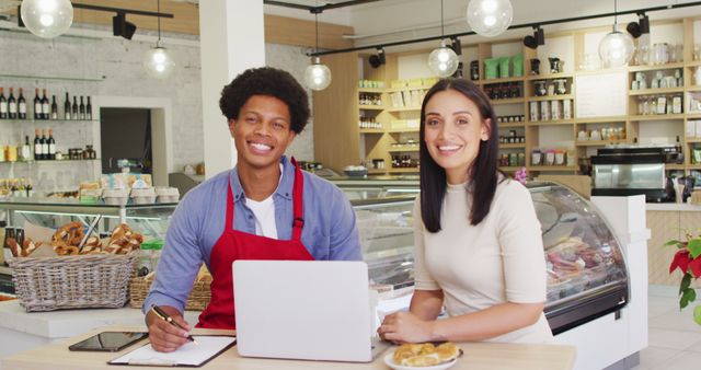 This image shows two cafe owners, a man and a woman, planning their business at the counter. They are using a laptop and taking notes while smiling at the camera. The background includes a well-organized coffee shop with a bakery counter and pastries. Ideal for use in articles or websites about small business management, entrepreneurship, cafes, teamwork, and hospitality industry.