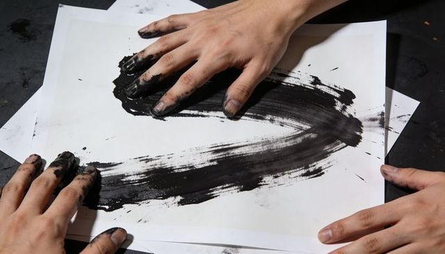 Three hands smearing black ink across paper creating bold abstract texture