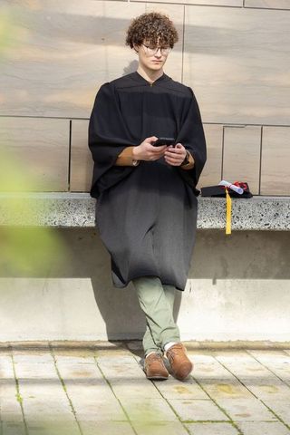 Graduating student checking smartphone while sitting on campus ledge with cap and tassel