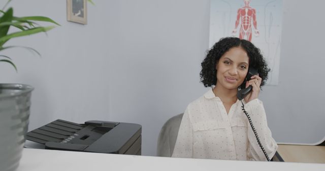 This image portrays a friendly receptionist answering the phone while seated at a desk in a professional office environment, suggesting a medical or healthcare setting. Use this for illustrating customer service, office jobs, or medical office settings, suitable for websites, brochures, and articles related to professional healthcare services, customer relations, and administrative work environments.