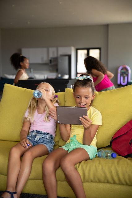 Two elementary-aged girls are sitting on a yellow couch in a play room. One girl is using a digital tablet while the other drinks water. In the background, other children are playing. This image can be used for educational content, technology in education, childhood development, and leisure activities.
