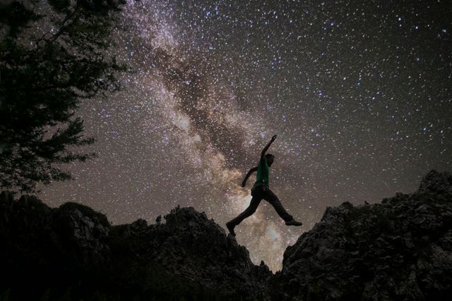Silhouette of man jumping over rocky terrain under the starry sky with the Milky Way. Perfect for themes related to outdoor adventure, space exploration, night photography, nature, and the awe of the universe. Suitable for promotional content for adventure gear, nature documentaries, astronomy events, motivational posters, and travel brochures.