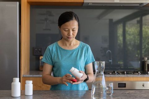 Senior asian woman reading supplement label at modern kitchen counter with glass of water