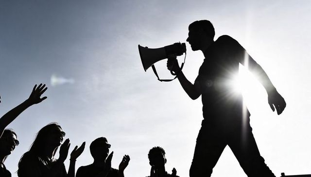 Speaking through megaphone leading silhouetted protest crowd under dramatic sunflare