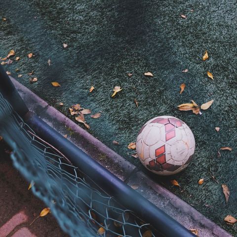 Worn soccer ball resting on turf beside chain-link fence with fallen leaves