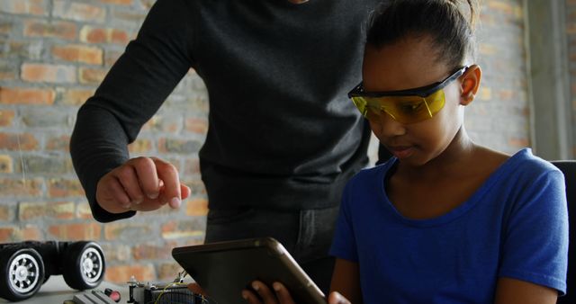Young African American girl learning electronics with assistance of her teacher. Both are focused on a tablet while the girl wears safety glasses. Ideal for illustrating concepts in STEM education, technology workshops, engineering classes, mentorship programs, and the importance of diversity in technology fields. Use for educational purposes, promotional materials for STEM programs, and ads highlighting mentoring and learning experiences.