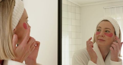 Woman applying pink under-eye hydrogel patches at mirror wearing white robe and headband