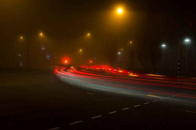 Long exposure capturing light trails of vehicles moving on foggy street corner at night. Suitable for themes of urban life, traffic, speed, and mystery, or illustrating concepts of night time movement and city glow.