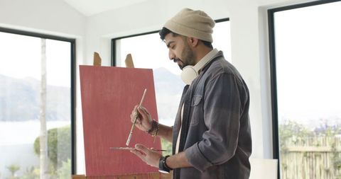 Indian male painter painting red canvas in bright studio with large windows and water view