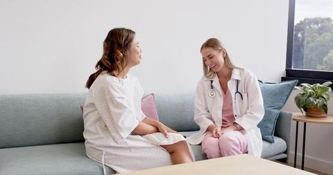 Diverse female doctor consulting with female patient on sofa in bright clinic room