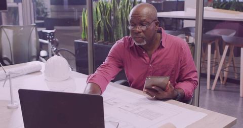 Senior Architect Analyzing Blueprints at Office Desk with Laptop