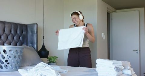 Smiling woman folding fresh towels while wearing headphones with laundry basket in cozy bedroom