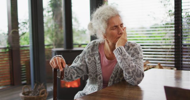 Senior woman wearing casual clothing yawning while sitting at a wooden table in a cozy room with natural light streaming through the windows. She holds onto a cane with one hand. The indoor setting features a calm, relaxed ambiance with a view of the greenery outside. This image can be used for themes of aging, relaxation, retirement, and daily life of seniors.