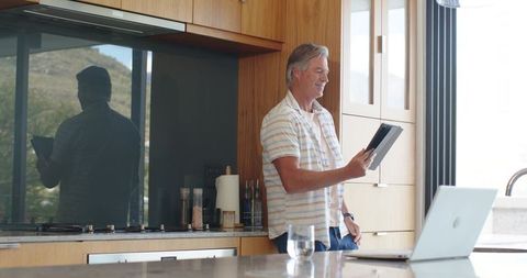 Middle-aged man checking tablet while using laptop in modern sunlit kitchen workspace