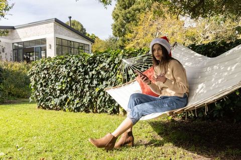 Relaxing woman wearing santa hat on hammock holding smartphone, gesturing outdoors
