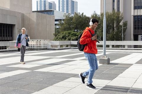 Student walking across campus plaza wearing red hoodie and headphones, holding smartphone