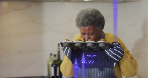 Senior woman smelling freshly baked muffins in apron and oven mitts with purple overlay