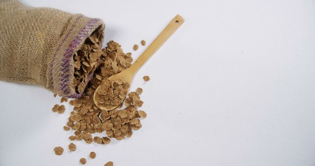 A burlap sack is tipped over, spilling crunchy cereal flakes onto a white surface, with a wooden spoon, with copy space. Ideal for showcasing natural and organic breakfast options or highlighting healthy eating concepts.
