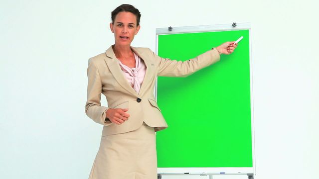 Businesswoman in a beige suit is gesturing and explaining in front of a green screen flip chart. Neutral background makes it ideal for text or graphics overlay about corporate communication, training sessions, or educational seminars.