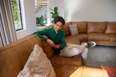 Young asian man relaxing on leather sofa using smartphone in bright modern living room
