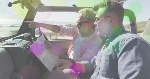 Two men pointing while driving convertible on coastal road holding map with sunlit bokeh