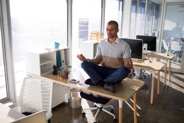 Male executive practicing yoga on desk in modern office, promoting mindfulness and relaxation in a corporate environment. Ideal for use in articles or advertisements about workplace wellness, stress relief, and work-life balance.