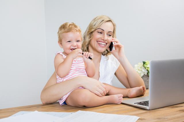 Mother holding baby girl while talking on mobile phone and using laptop at home. Ideal for themes related to parenting, work-life balance, remote work, family life, and modern motherhood.